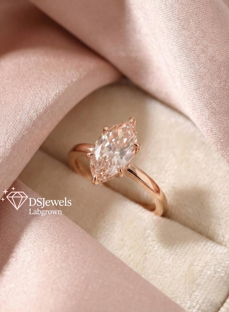 May include: A rose gold engagement ring with a marquise-cut lab-grown diamond. The diamond is a light peach color and set in a four-prong setting. The ring is displayed on a pink satin fabric. The text "DSJewels Labgrown" is visible.