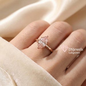 May include: A rose gold ring featuring a marquise-cut, lab-grown diamond. The diamond has a soft pink hue and is set on a delicate band. The ring is displayed on a hand against a cream-colored background. The text "DSJewels Labgrown" is visible.