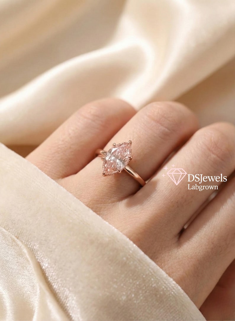 May include: A rose gold ring featuring a marquise-cut, lab-grown diamond. The diamond has a soft pink hue and is set on a delicate band. The ring is displayed on a hand against a cream-colored background. The text "DSJewels Labgrown" is visible.