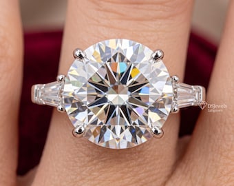 Può includere: Un anello color argento con una grande pietra centrale rotonda a taglio brillante fissata da sei griffe, affiancata da due pietre laterali a taglio baguette affusolato. Il gambo è in metallo lucido. Il testo DSjewels Labgrown è visibile a destra.
