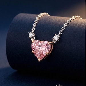 Puede incluir: Un delicado collar con un gran colgante de piedra preciosa rosa en forma de corazón. El colgante está engastado en un marco dorado y cuelga de una cadena plateada. Pequeñas piedras preciosas transparentes adornan la cadena. El fondo es azul oscuro.