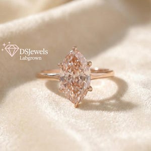 May include: A marquise-cut, lab-grown diamond ring with a rose gold band. The diamond has a light peach hue and is set with six prongs. The ring is displayed on a soft, off-white fabric. The logo "DSJewels Labgrown" is in the upper left corner.