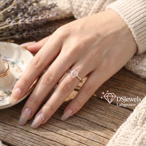 May include: A silver ring with a pear-shaped, light pink gemstone. The ring is on a hand with long, manicured nails. The background includes a wooden surface, a white sweater, and a sprig of lavender. The logo "DSJewels Labgrown" is in the bottom right corner.