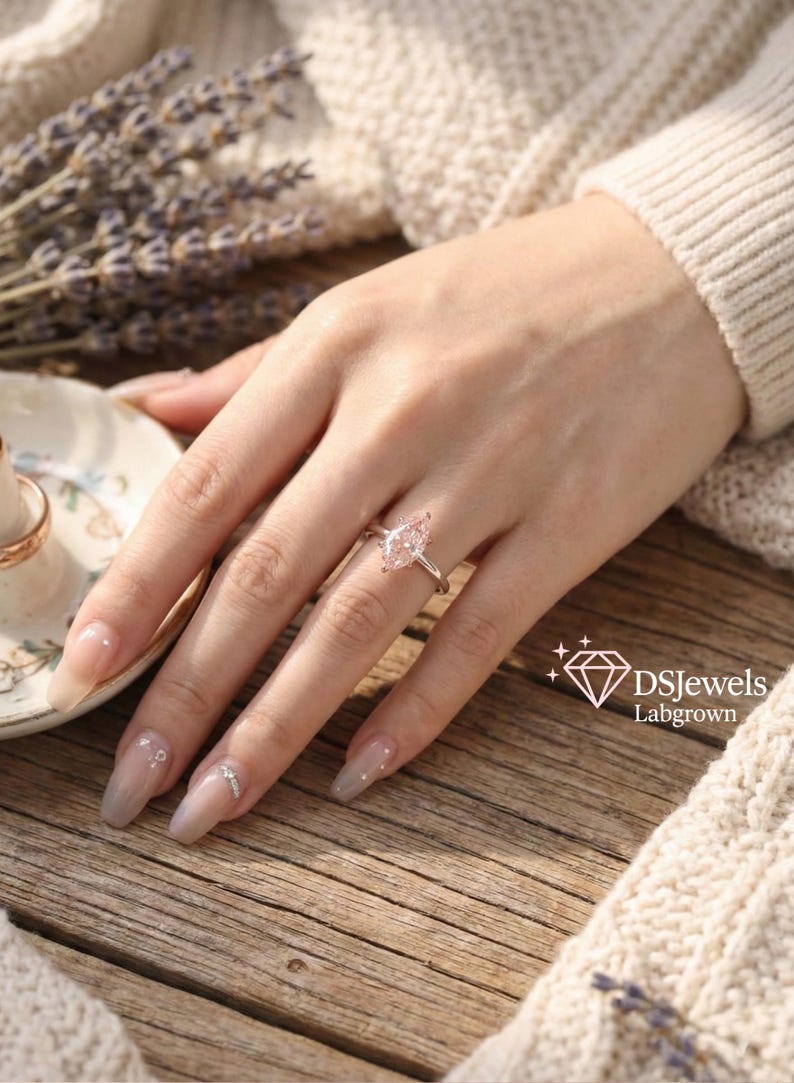 May include: A silver ring with a pear-shaped, light pink gemstone. The ring is on a hand with long, manicured nails. The background includes a wooden surface, a white sweater, and a sprig of lavender. The logo "DSJewels Labgrown" is in the bottom right corner.