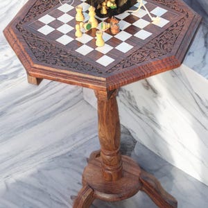 May include: A handcrafted wooden chess table with an octagonal top. The table features a carved floral design around the edges and a checkered playing surface. Chess pieces and a black velvet bag are on the table. The table has a single pedestal leg and three feet.