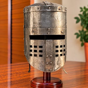 Op de afbeelding: Een cilindrische metalen helm met een verweerde, gepitte oppervlaktestructuur. De helm is voorzien van een horizontale kijkspleet en een onderste voorplaat met twee rechthoekige ventilatieopeningen in rasterpatroon, vastgezet met zichtbare klinknagels.