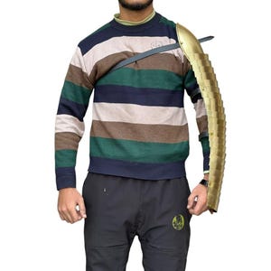 Op de afbeelding: Een goudkleurige armbeschermer met een segmentontwerp, vastgezet met een zwarte riem. Het pantser wordt gedragen over een gestreepte trui in tinten groen, bruin en marineblauw. De algehele esthetiek suggereert een historisch of fantasiethema.