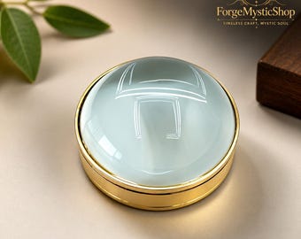 May include: A circular paperweight featuring a smooth, pale blue glass dome set within a polished gold-tone metal base. The object has a reflective surface and a clean, minimalist design. The ForgeMysticShop logo is visible in the upper right corner.