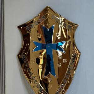 Op de afbeelding: Een goudkleurig schild met een blauw kruis in het midden. Het schild heeft een decoratieve rand en is bevestigd aan een lichtgrijze muur. Het ontwerp is in middeleeuwse stijl.