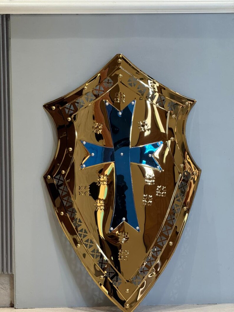 Op de afbeelding: Een goudkleurig schild met een blauw kruis in het midden. Het schild heeft een decoratieve rand en is bevestigd aan een lichtgrijze muur. Het ontwerp is in middeleeuwse stijl.