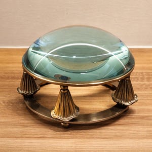 May include: A large, clear magnifying glass with a teal tint, set on a brass and metal stand. The stand has three ornate, cone-shaped legs and a circular base. The magnifying glass is likely used for reading or detailed work.
