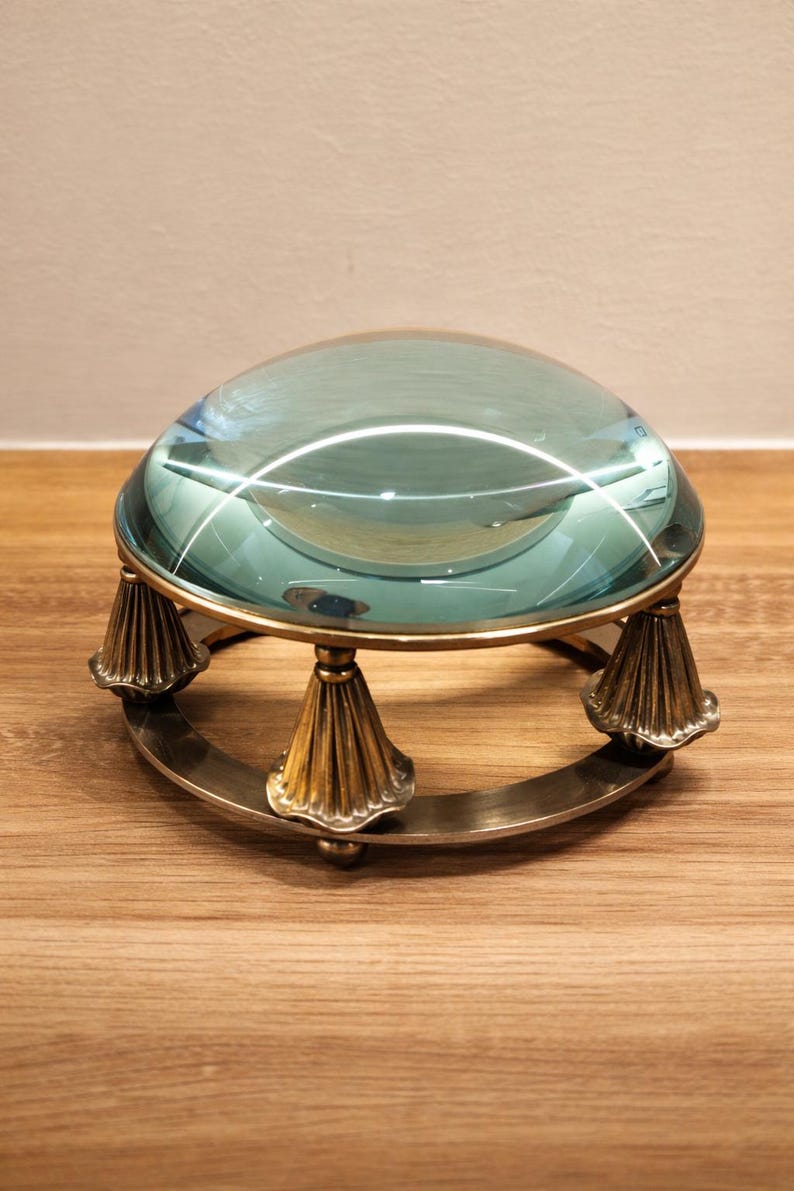 May include: A large, clear magnifying glass with a teal tint, set on a brass and metal stand. The stand has three ornate, cone-shaped legs and a circular base. The magnifying glass is likely used for reading or detailed work.
