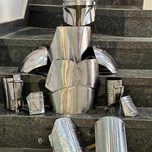 May include: A complete set of polished metal armor, including a helmet, chest plate, shoulder guards, arm guards, and leg guards. The armor is displayed on a set of stairs, showcasing its intricate design and reflective surface.
