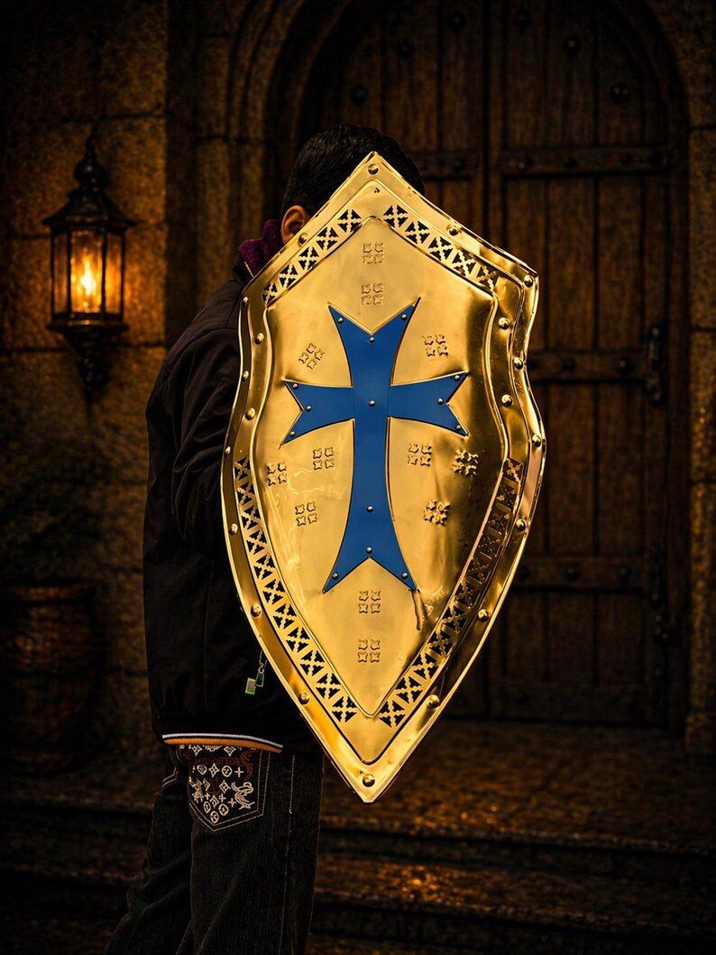 Op de afbeelding: Een goudkleurig schild met een blauw kruisontwerp. Het schild heeft een decoratieve rand. De achtergrond toont een stenen muur, een houten deur en een lantaarn. Het schild wordt voor een persoon gehouden.