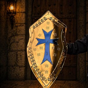 Op de afbeelding: Een goudkleurig schild met een blauw kruisontwerp. Het schild heeft een decoratieve rand en wordt gehouden tegen een vage achtergrond van een stenen muur en houten deur.