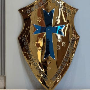 Op de afbeelding: Een goudkleurig schild met een blauw kruis in het midden. Het schild heeft een decoratieve rand en een reflecterend oppervlak. Het is een item met een middeleeuws thema.