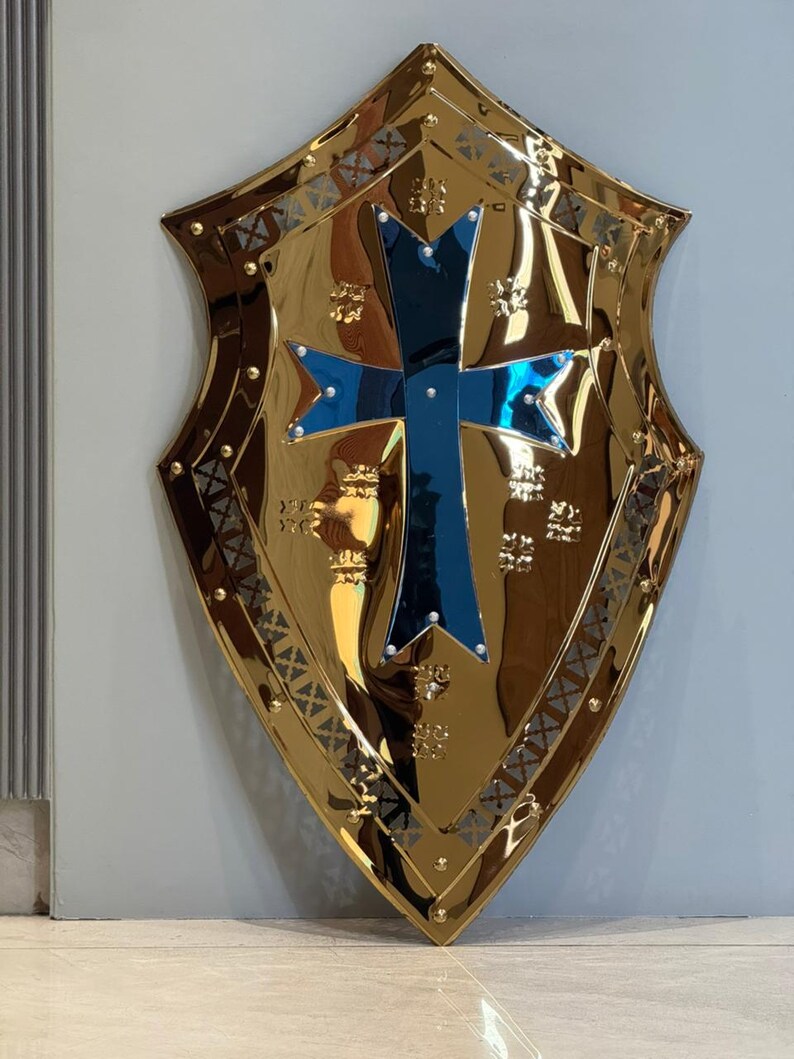 Op de afbeelding: Een goudkleurig schild met een blauw kruis in het midden. Het schild heeft een decoratieve rand en een reflecterend oppervlak. Het is een item met een middeleeuws thema.