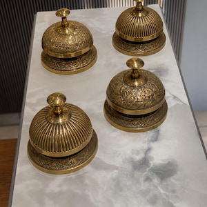 Peut inclure: Quatre sonnettes de comptoir en laiton dispos&eacute;es en carr&eacute;. Deux sonnettes pr&eacute;sentent un motif floral en relief sur le d&ocirc;me, tandis que les deux autres ont une texture nervur&eacute;e verticale. Chaque sonnette poss&egrave;de une base ronde et un bouton.