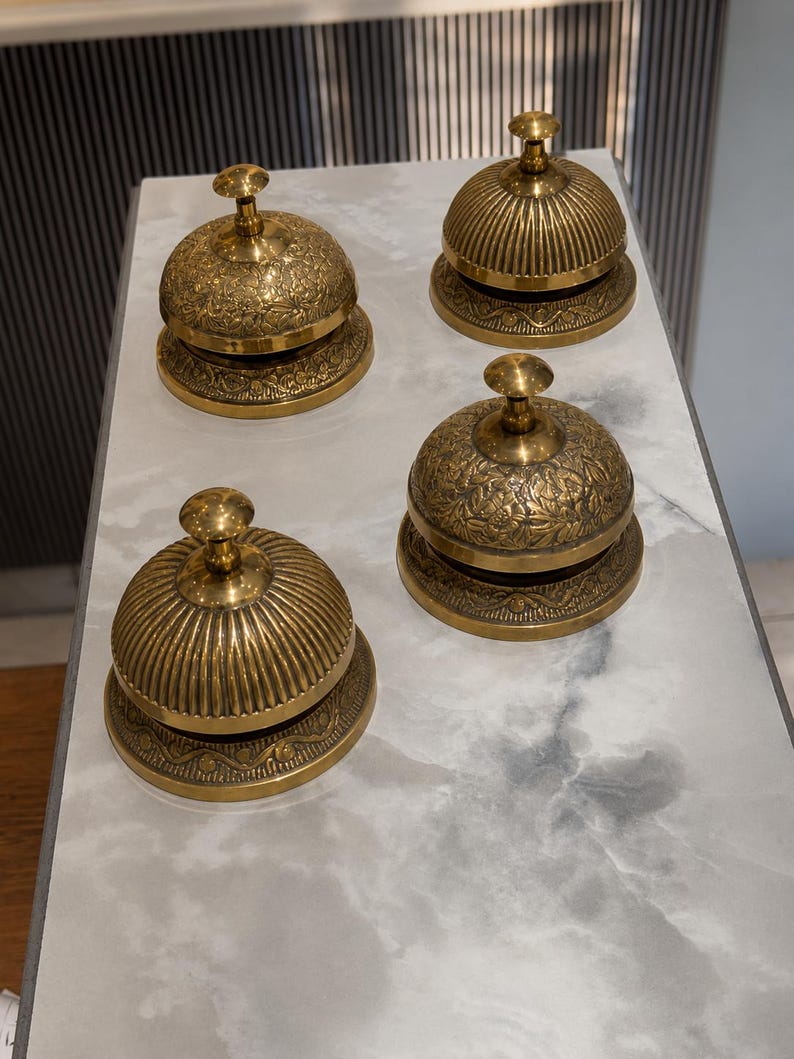 Peut inclure: Quatre sonnettes de comptoir en laiton dispos&eacute;es en carr&eacute;. Deux sonnettes pr&eacute;sentent un motif floral en relief sur le d&ocirc;me, tandis que les deux autres ont une texture nervur&eacute;e verticale. Chaque sonnette poss&egrave;de une base ronde et un bouton.