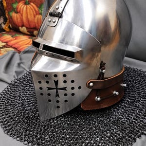 Op de afbeelding: Een gepolijste stalen ridderhelm met een scharnierend vizier met een horizontale oogopening en een onderste gezichtsbeschermer met een kruisvormige uitsparing omringd door cirkelvormige ventilatiegaten en een bruine leren nekband met klinknagels.