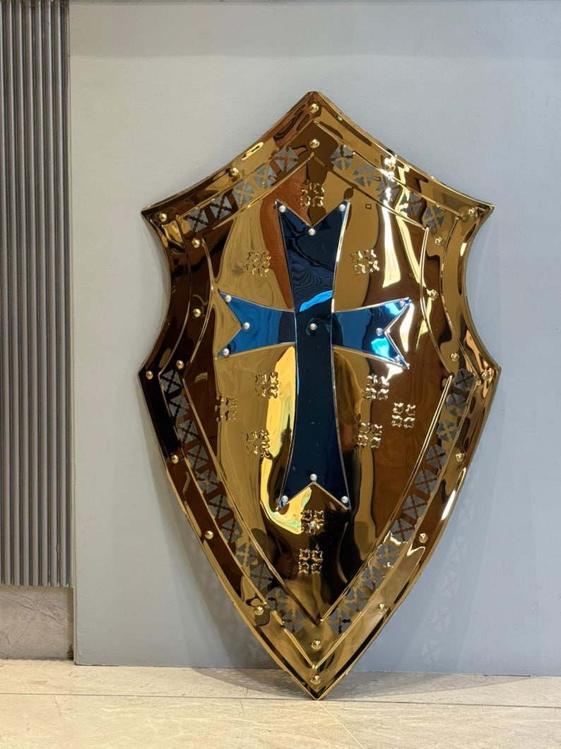 Op de afbeelding: Een goudkleurig schild met een centraal kruisontwerp. Het schild heeft een reflecterend oppervlak en is versierd met decoratieve patronen langs de randen en rond het kruis. Het kruis is zwart met blauwe accenten.