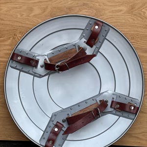 K&ouml;nnte beinhalten: Ein silberfarbener, runder Schild mit konzentrischen Kreisen und zwei grauen Armst&uuml;tzen. Die St&uuml;tzen haben braune Lederriemen mit silbernen Schnallen und Nieten. Der Schild wird auf einer Holzoberfl&auml;che pr&auml;sentiert.