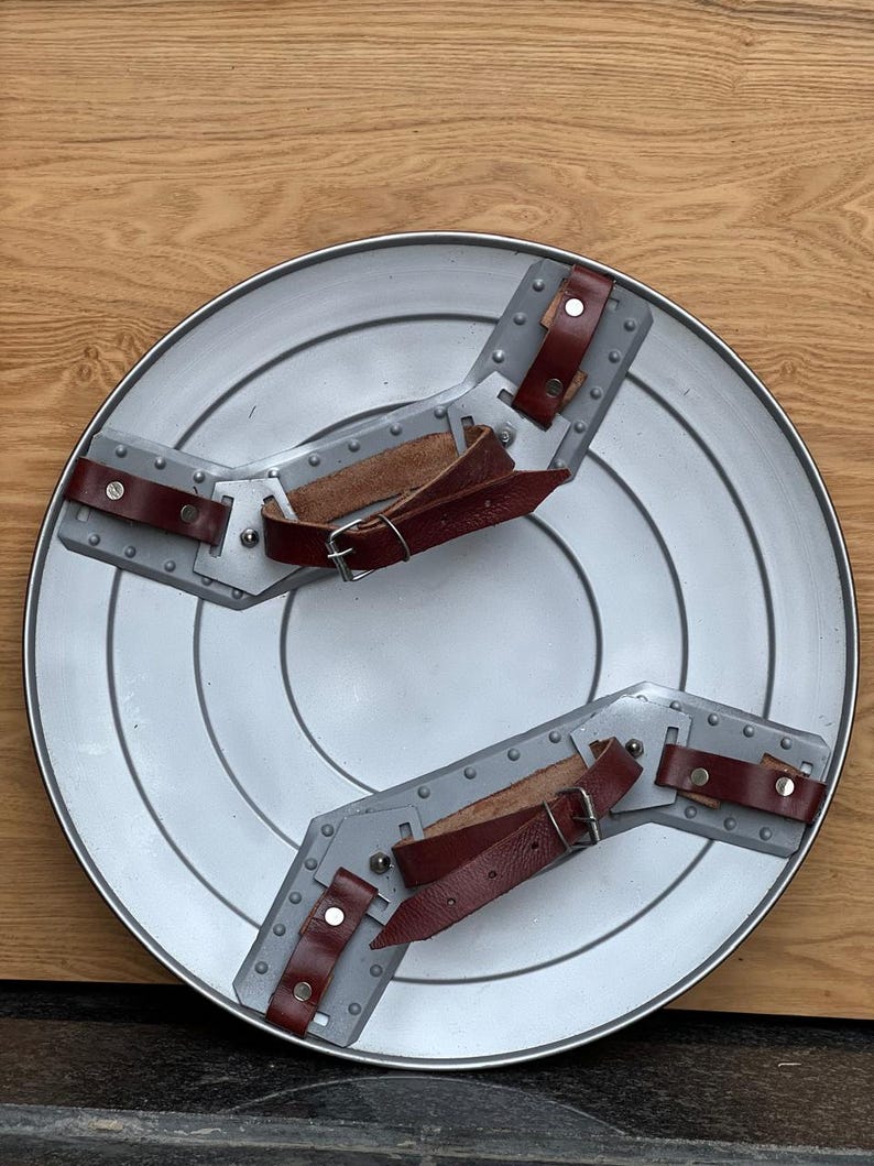 K&ouml;nnte beinhalten: Ein silberfarbener, runder Schild mit konzentrischen Kreisen und zwei grauen Armst&uuml;tzen. Die St&uuml;tzen haben braune Lederriemen mit silbernen Schnallen und Nieten. Der Schild wird auf einer Holzoberfl&auml;che pr&auml;sentiert.