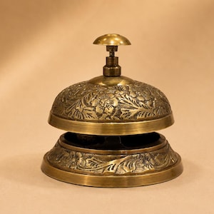 Peut inclure: Une sonnette de bureau en laiton avec un d&ocirc;me arrondi et une base circulaire. La surface ext&eacute;rieure de la cloche et de la base pr&eacute;sente un motif floral en relief avec des feuilles et des fleurs. Un petit bouton arrondi lisse se trouve au sommet.