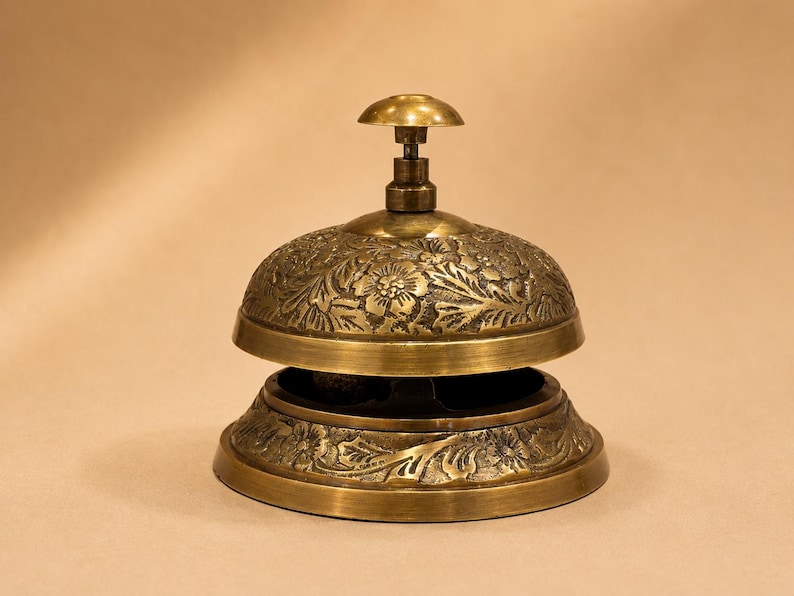 Peut inclure: Une sonnette de bureau en laiton avec un d&ocirc;me arrondi et une base circulaire. La surface ext&eacute;rieure de la cloche et de la base pr&eacute;sente un motif floral en relief avec des feuilles et des fleurs. Un petit bouton arrondi lisse se trouve au sommet.