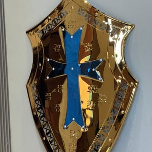 Op de afbeelding: Een goudkleurig schild met een blauw kruisontwerp in het midden. Het schild heeft een reflecterend oppervlak en is versierd met ingewikkelde patronen en klinknagels langs de randen. Het kruis heeft witte accenten.