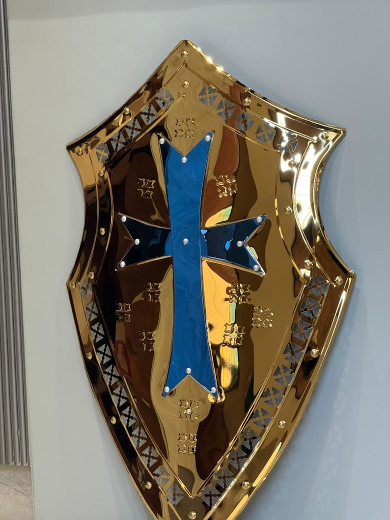 Op de afbeelding: Een goudkleurig schild met een blauw kruisontwerp in het midden. Het schild heeft een reflecterend oppervlak en is versierd met ingewikkelde patronen en klinknagels langs de randen. Het kruis heeft witte accenten.