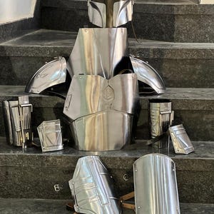 May include: A complete set of polished metal armor, including a helmet, chest plate, shoulder guards, and various other protective pieces. The armor is displayed on a set of stairs, showcasing its intricate design and reflective surface.