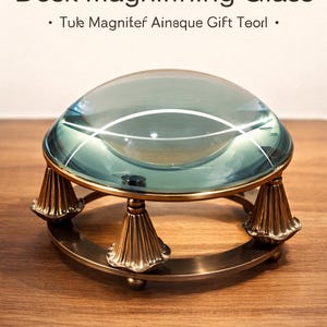 May include: Vintage brass desk magnifying glass with a clear, rounded lens. The magnifying glass is supported by a brass base with decorative, cone-shaped feet. The text "Vintage Brass Desk Magnifying Glass" is at the top.