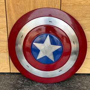 K&ouml;nnte beinhalten: Ein runder Schild im Captain America-Design. Er hat einen roten Au&szlig;enring, einen silbernen Ring und eine blaue Mitte mit einem wei&szlig;en Stern. Der Schild ist eine Nachbildung.