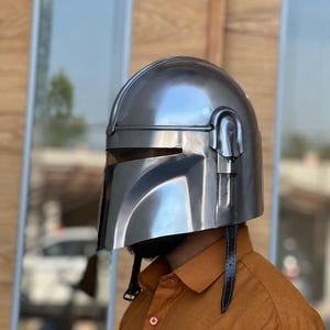 May include: A silver helmet with a visor and a black leather strap. The helmet has a polished, metallic finish and is designed with angular features. The helmet is worn by a person wearing an orange button-down shirt.