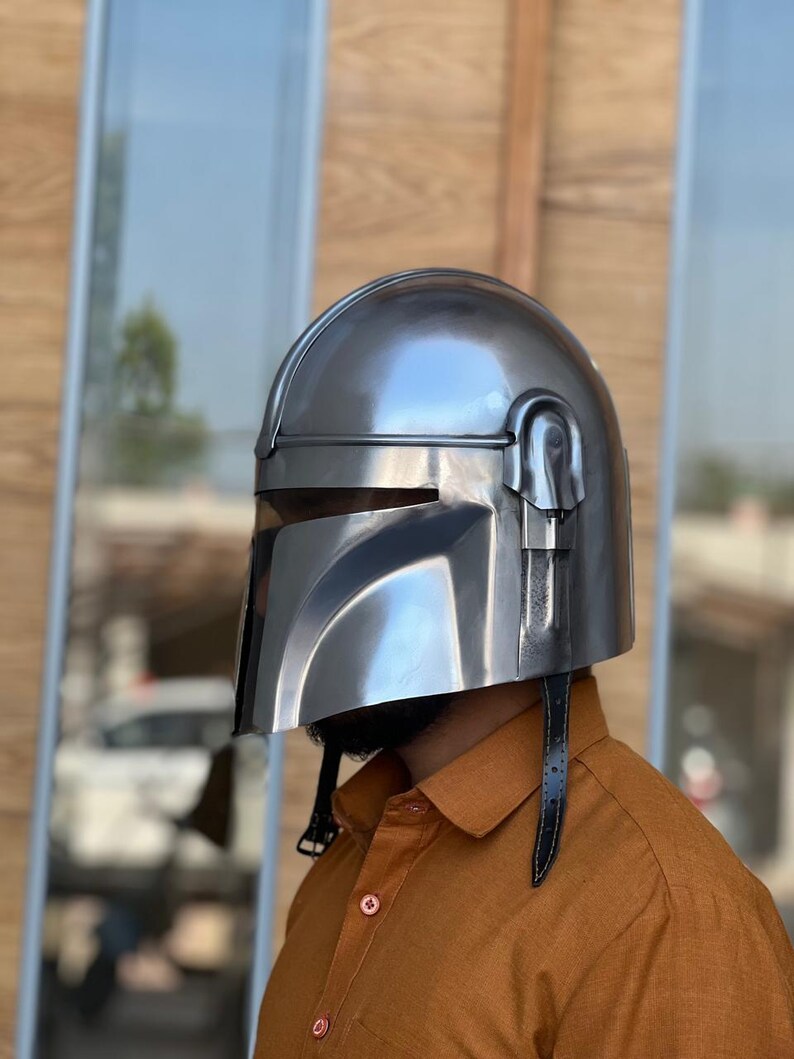 May include: A silver helmet with a visor and a black leather strap. The helmet has a polished, metallic finish and is designed with angular features. The helmet is worn by a person wearing an orange button-down shirt.