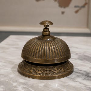 Peut inclure: Une cloche de r&eacute;ception en laiton de style ancien avec un sommet en forme de d&ocirc;me pr&eacute;sentant des cannelures verticales. La base est orn&eacute;e d'un motif de volutes r&eacute;p&eacute;titif en relief. Le m&eacute;tal a une patine sombre et vieillie avec un petit bouton.