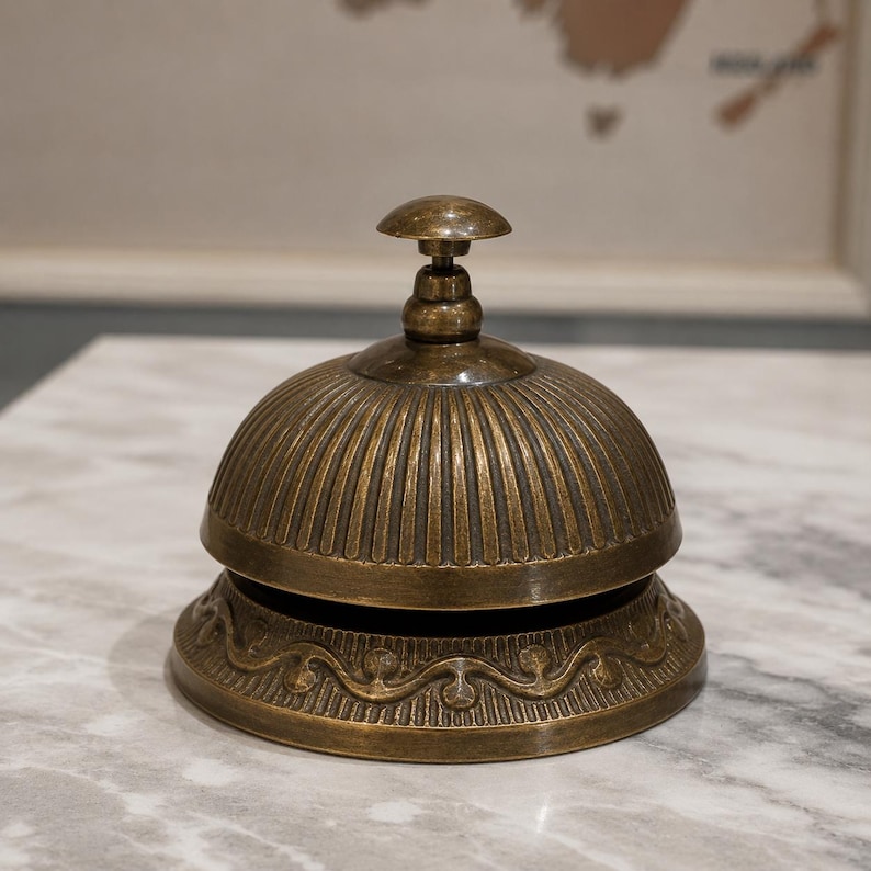 Peut inclure: Une cloche de r&eacute;ception en laiton de style ancien avec un sommet en forme de d&ocirc;me pr&eacute;sentant des cannelures verticales. La base est orn&eacute;e d'un motif de volutes r&eacute;p&eacute;titif en relief. Le m&eacute;tal a une patine sombre et vieillie avec un petit bouton.