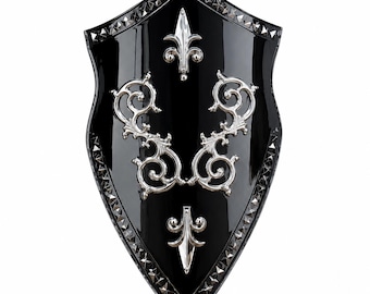 May include: A black shield with a glossy finish featuring a silver-toned border of small pyramid studs. The center is decorated with two silver-toned fleur-de-lis emblems and two symmetrical silver-toned scrollwork ornaments arranged vertically.