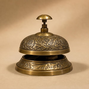 Peut inclure: Une cloche de r&eacute;ception en laiton orn&eacute;e d'un motif floral en relief sur le d&ocirc;me et la base. La cloche pr&eacute;sente un bouton sup&eacute;rieur arrondi et lisse pour l'activation, avec une finition m&eacute;tallique polie et des d&eacute;tails sombres en creux.