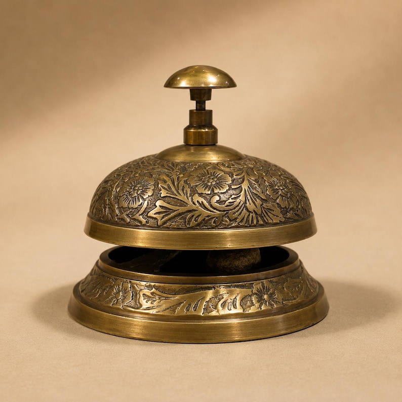 Peut inclure: Une cloche de r&eacute;ception en laiton orn&eacute;e d'un motif floral en relief sur le d&ocirc;me et la base. La cloche pr&eacute;sente un bouton sup&eacute;rieur arrondi et lisse pour l'activation, avec une finition m&eacute;tallique polie et des d&eacute;tails sombres en creux.