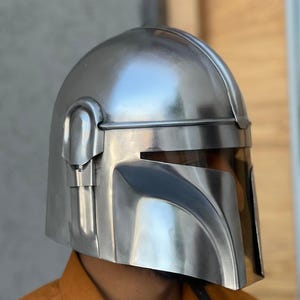 May include: A silver Mandalorian helmet with a visor. The helmet has a smooth, metallic finish and is designed with angular features. The visor is tinted, and the helmet appears to be a replica or a costume piece.