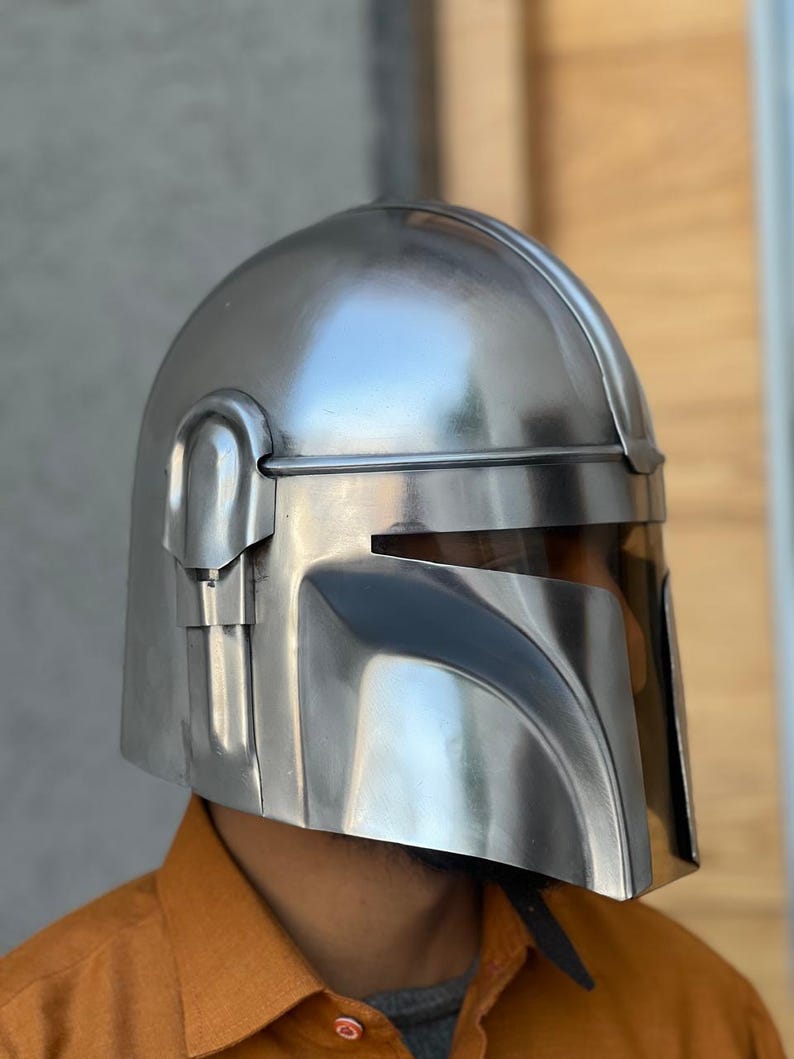 May include: A silver Mandalorian helmet with a visor. The helmet has a smooth, metallic finish and is designed with angular features. The visor is tinted, and the helmet appears to be a replica or a costume piece.