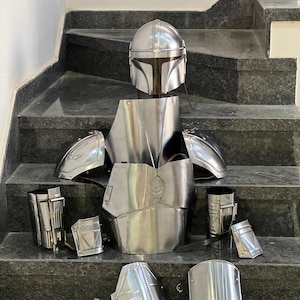 May include: A complete set of silver-colored Mandalorian armor, including a helmet, chest plate, shoulder pads, and various leg and arm pieces. The armor is displayed on a set of stairs.