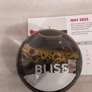 May include: A round paperweight with a clear glass dome. The paperweight features a field of sunflowers and the word "BLISS" in white. A calendar page with the text "MAY 2025" is in the background.