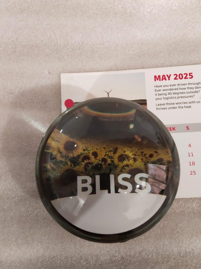 May include: A round paperweight with a clear glass dome. The paperweight features a field of sunflowers and the word "BLISS" in white. A calendar page with the text "MAY 2025" is in the background.