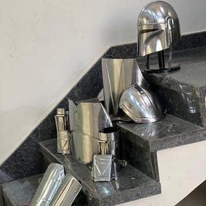 May include: A collection of shiny, silver-colored metal armor pieces, including a helmet, chest plate, and various arm and leg guards. The helmet is displayed on a black stand. The armor is arranged on a set of gray marble steps.