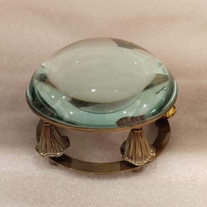May include: A clear glass magnifying lens with a brass base. The lens is dome-shaped and sits atop a decorative brass stand with a curved base and ornate, fan-shaped supports. The glass has a slight green tint.