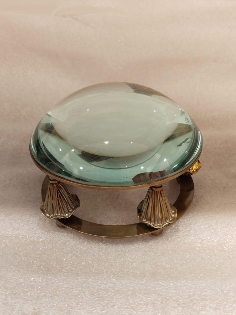 May include: A clear glass magnifying lens with a brass base. The lens is dome-shaped and sits atop a decorative brass stand with a curved base and ornate, fan-shaped supports. The glass has a slight green tint.