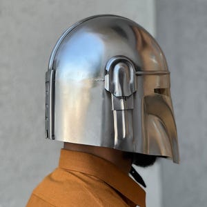 May include: A silver helmet with a unique visor and rounded crown. The metallic helmet has a reflective surface. The design includes vertical lines on the side and a curved visor opening. The helmet is likely made of metal.